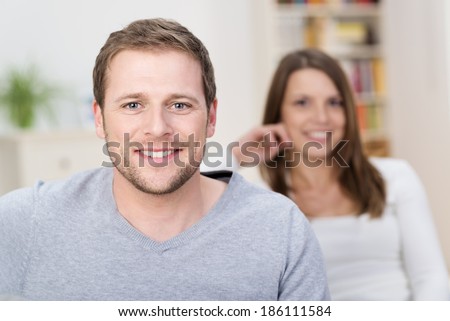 Handsome young man with a friendly smile sitting in the living room smiling at the camera watched by hid wife behind, focus to his face