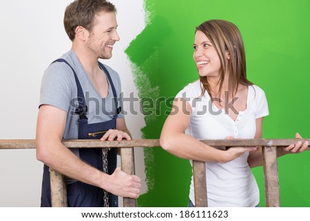 Happy young couple laughing together as they redecorate their new home and work as a team carrying a wooden stepladder