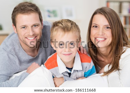 Happy little boy with his smiling young parents posing together in their home sitting on a sofa looking at the camera with warm friendly smiles