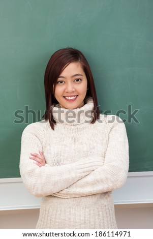 Confident young Asian student or teacher in a stylish polo-neck sweater standing in front of the class blackboard with folded arms