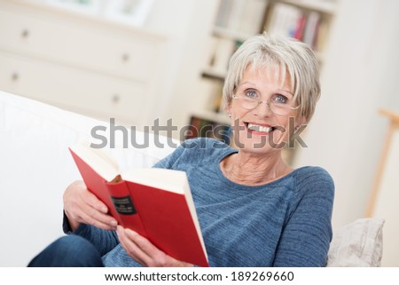 Smiling friendly senior woman sitting on a couch in her living room at home reading a book and looking up at the camera with a vivacious smile