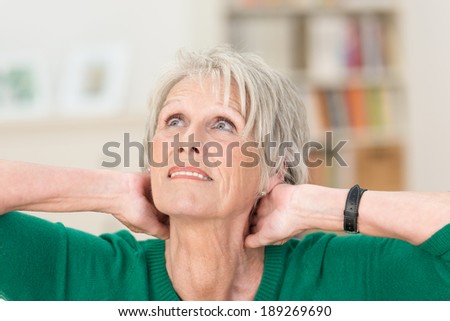 Senior women sitting thinking in her living room with her hands clasped behind her neck staring up to heaven with a concerned expression