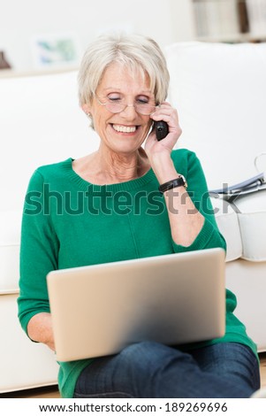 Senior lady chatting on her mobile phone while relaxing on the floor in her living room working on a laptop computer smiling as she listens to the conversation