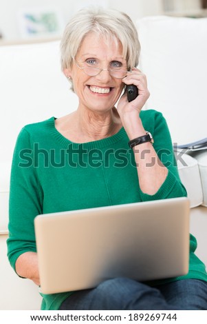 Delighted attractive elderly woman chatting on a mobile phone with her laptop balanced on her knee smiling happily at the camera