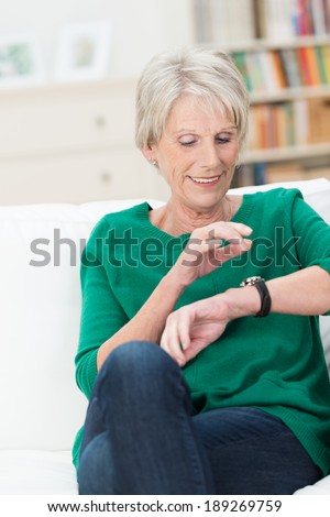 Beautiful elderly woman checking the time looking at her wrist watch as she relaxes on a sofa in her living room