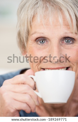Elderly woman enjoying a relaxing cup of tea smiling over the rim of the cup as she looks directly into the lens