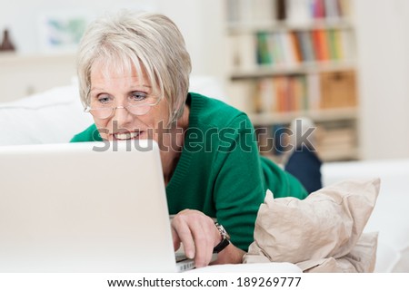 Tech savvy senior woman relaxing with a laptop at home lying on a sofa wearing her glasses and reading the screen
