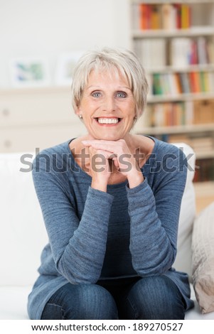 Beautiful trendy vivacious elderly woman sitting on a sofa in the living room beaming happily at the camera