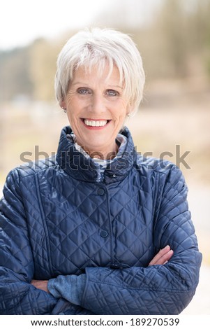 Fashionable attractive senior woman in a high necked jacket standing outdoors with folded arms smiling at the camera