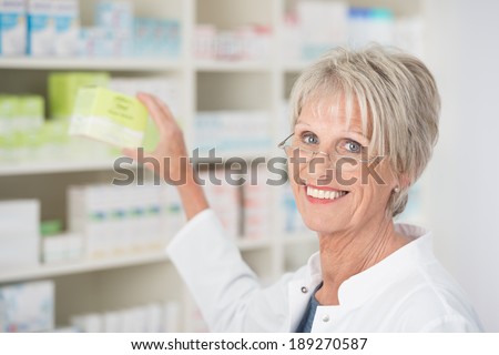 Smiling happy female pharmacist reaching for medication on the shelf and displaying it to the camera