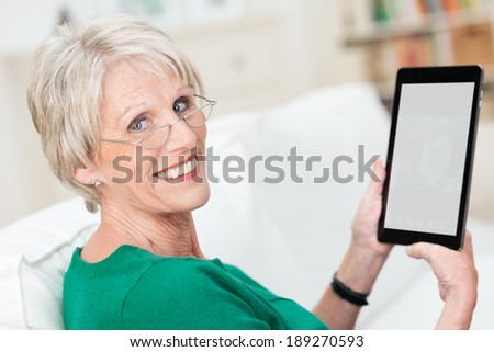 Attractive senior woman using a tablet computer relaxing on a sofa at home turning to smile at the camera - the screen blank is visible to the viewer