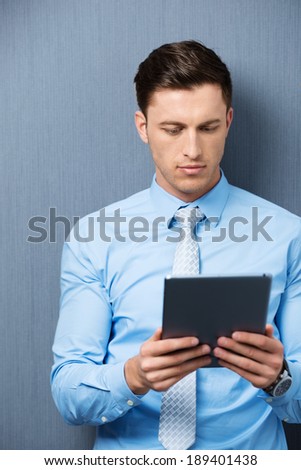 Businessman standing against a green background reading information on his handheld tablet computer with a serious expression