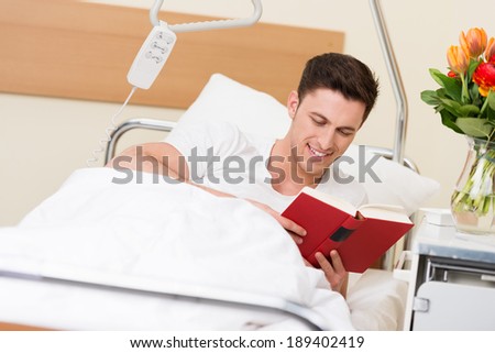 Young man recuperating from an illness or injury in hospital lying reading a book with a smile of enjoyment