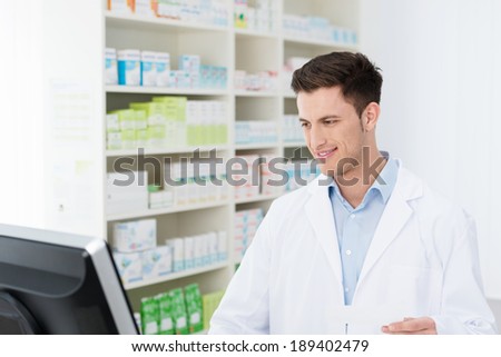 Smiling young male pharmacist standing checking stock on his computer in the pharmacy as he prepares to dispense medication