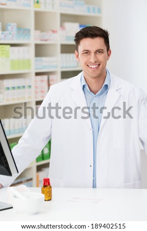 Handsome young pharmacist at work in the pharmacy standing behind the counter dispensing medicines and smiling at the camera