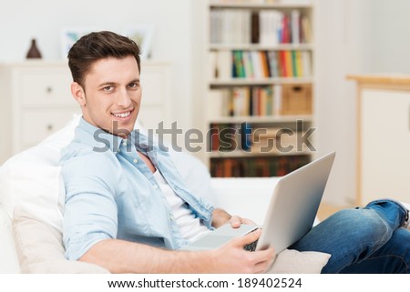 Young man relaxing at home with a laptop computer on his lap as he sits on a comfortable sofa in the living room smiling at the camera
