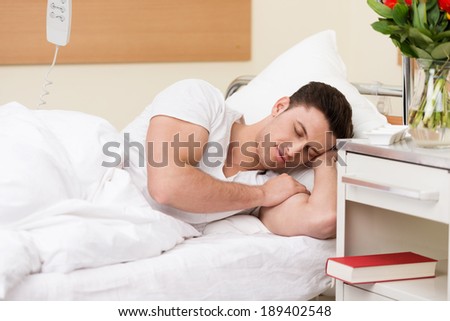 Young man having a restful sleep in a hospital bed with his book on the night stand alongside him as he recuperates from an illness or injury