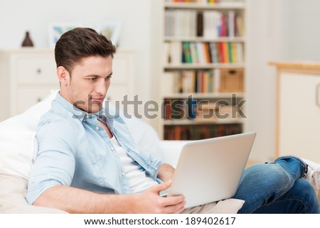 Young man concentrating as he works on a laptop at home as he relaxes on a sofa in the living room