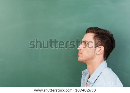 Thoughtful young man in a classroom standing in profile in front of a blank green blackboard with copyspace staring pensively into the distance