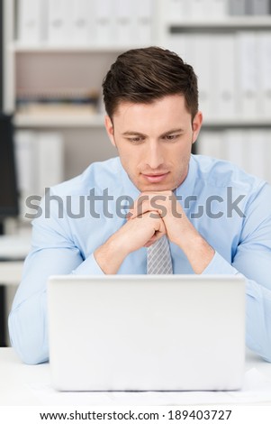 Thoughtful young businessman sitting at his desk reading his laptop screen with his chin resting on his hands as he concentrates on the information, frontal view