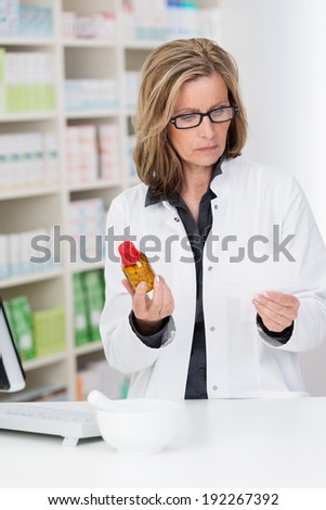 Attractive middle-aged female pharmacist checking a prescription against a bottle of tablets in her hand as she stands working at the counter in the pharmacy