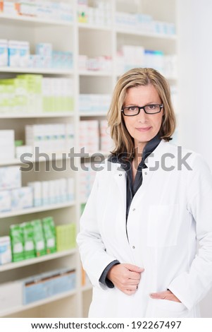 Attractive friendly woman pharmacist wearing glasses standing with her hand in the pocket of her white coat against stocked shelves smiling at the camera