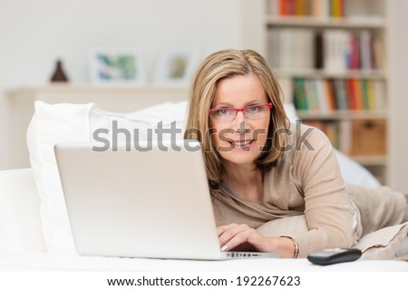 Middle-aged woman relaxing at home with her laptop computer lying on her stomach on a sofa smiling at the camera