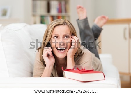 Attractive middle-aged woman lying on her stomach on a sofa laughing as she chats on her mobile phone