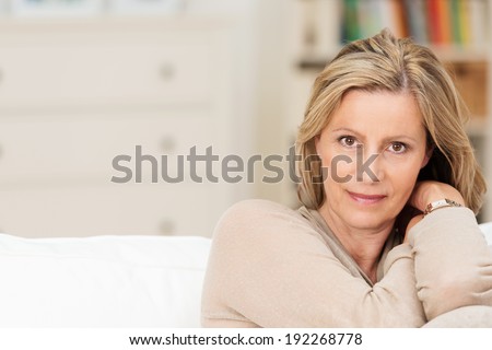 Attractive sincere middle-aged woman sitting on a sofa leaning her head on her raised arm looking directly at the camera with a serious expression