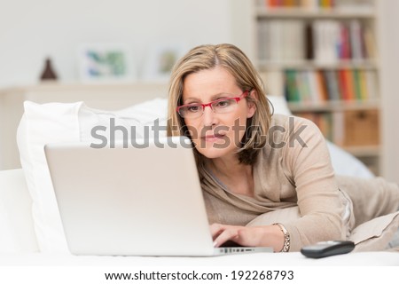 Woman wearing glasses lying on a sofa at home concentrating as she works on a laptop