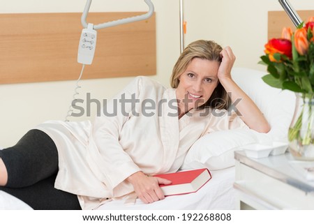 Smiling middle-aged attractive woman lying on a hospital bed holding a closed book in her hand as she recuperates from an illness or injury