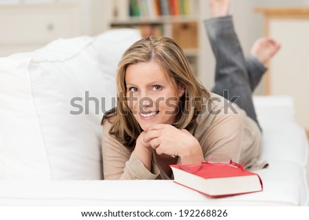 Happy relaxed woman with her book lying on her stomach on a sofa in the living room smiling at the camera