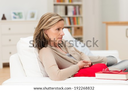 Attractive middle-aged depressed worried woman relaxing at home lying on a sofa staring straight ahead with a serious expression