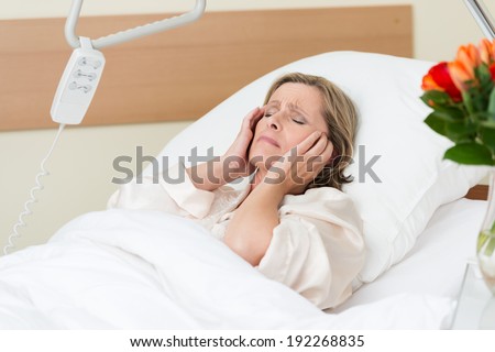 Woman in pain in a hospital bed clutching at her temples as she grimaces in agony from a migraine or head pain as she reclines back against the fresh white pillows