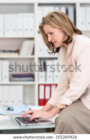 Businesswoman perched on the edge of her desk in the office typing in information on her laptop computer, side view against shelves of binders