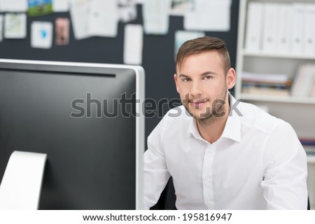 Young businessman sitting at his desk in the office in front of a large desktop computer looking at the camera with a smile