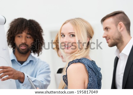 Smiling attractive blond businesswoman turning to look at the camera as she has a discussion with two multiethnic male colleagues