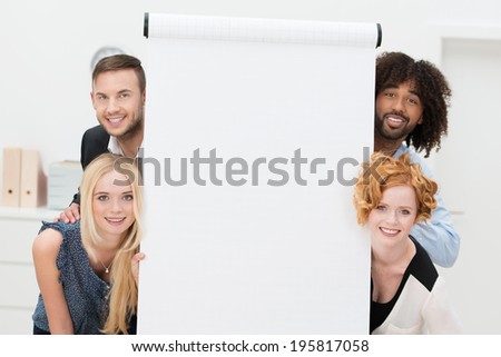 Smiling successful multiethnic business team with a blank white sheet of paper on a flip chart peering playfully around the sides at the camera