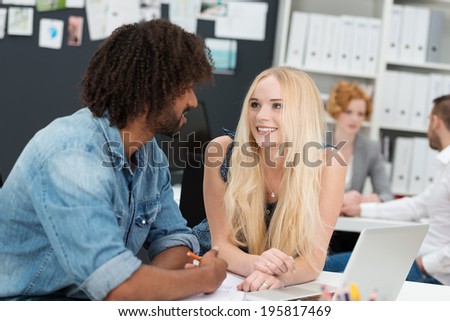 Friendly pretty young businesswoman chatting to a male African American colleague as they sit working at a desk together in the office
