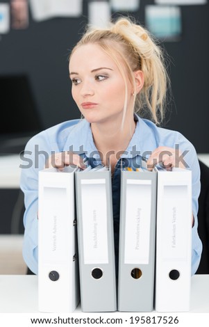 Pensive young businesswoman with a heavy workload and row of large files staring thoughtfully off to the side