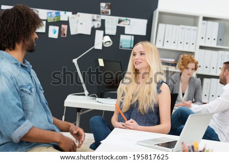 Two young businesspeople in a busy office sitting chatting with a beautiful young blond woman and young African American male in the foreground and colleagues working behind