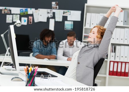 Happy beautiful contented young businesswoman relaxing in her chair stretching her arms in the air in a busy office with multiethnic male business partners