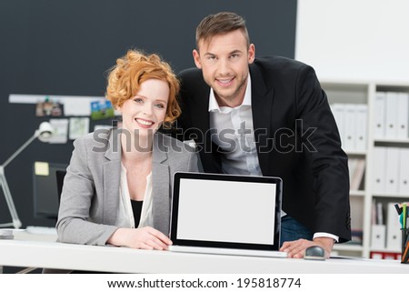 Two attractive smiling businesspeople displaying their team project on a blank tablet computer standing upright on a desk visible to the viewer