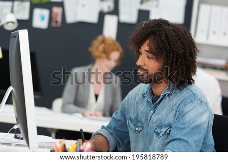 Casual young African American businessman in a denim shirt sitting working st his computer in the office with a quiet smile of satisfaction