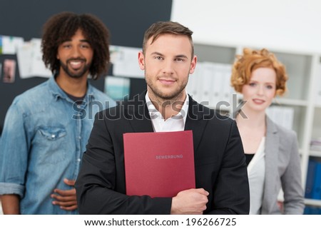 Successful male job appplicant standing in a suit holding a file with his CV flanked by two mutliethnic colleagues. German word BEWERBUNG means application