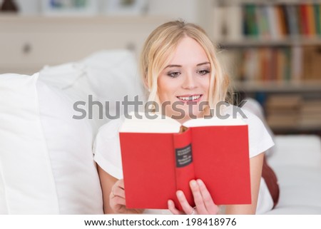Beautiful woman relaxing with a good book on the sofa at home smiling with enjoyment as she reads the story