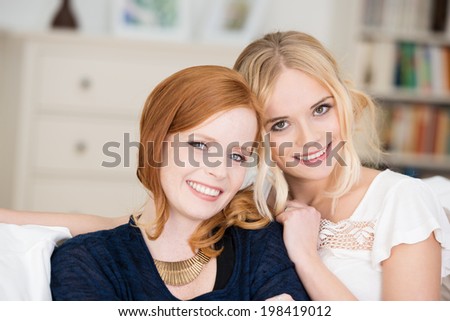 Two beautiful women friends posing with their heads close together in a living room looking at the camera with charming smiles