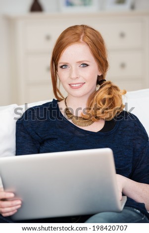 Beautiful young woman working at home sitting on a sofa with a laptop computer on her lap smiling at the camera