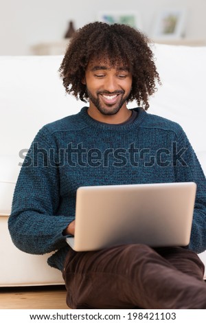 Handsome hip young African American man smiling as he types on his laptop computer while relaxing on the floor in the living room
