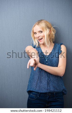 Laughing attractive young woman standing against a grey background pointing at the camera as she identifies someone or singles out her choice